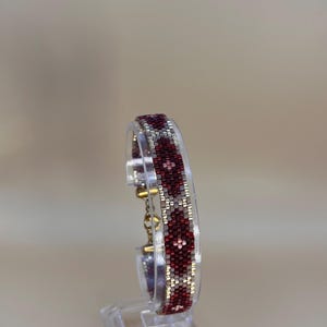 May include: A beaded bracelet with a geometric pattern in shades of burgundy, silver, and gold. The bracelet is displayed on a clear acrylic stand, showcasing its intricate design and craftsmanship. The bracelet's pattern is a repeating series of diamond shapes.
