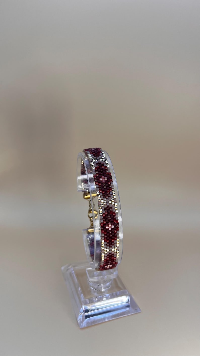 May include: A beaded bracelet with a geometric pattern in shades of burgundy, silver, and gold. The bracelet is displayed on a clear acrylic stand, showcasing its intricate design and craftsmanship. The bracelet's pattern is a repeating series of diamond shapes.