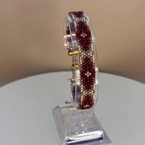 May include: A beaded bracelet with a geometric pattern in shades of red, gold, and white. The bracelet is displayed on a clear acrylic stand, showcasing its intricate design and craftsmanship. The bracelet is a stylish accessory.
