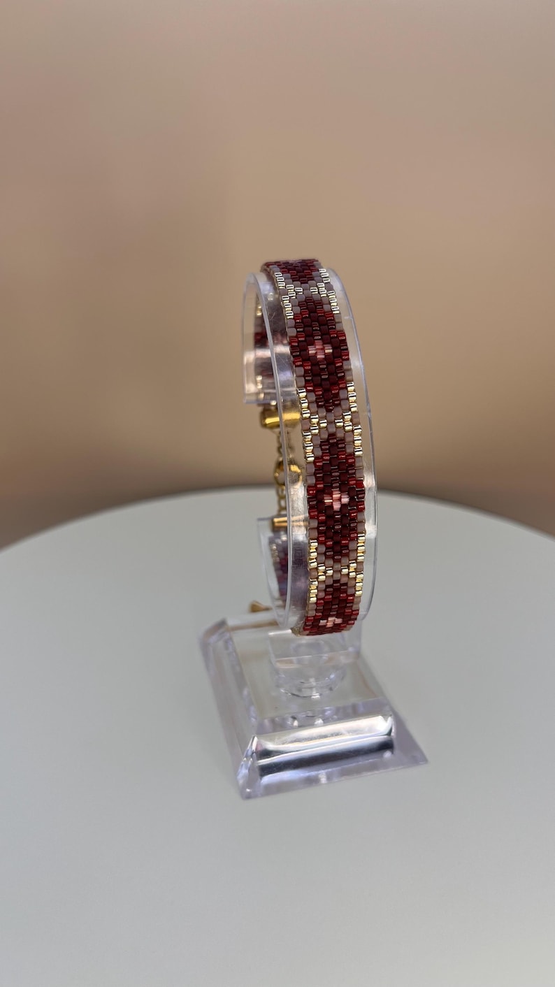 May include: A beaded bracelet with a geometric pattern in shades of red, gold, and white. The bracelet is displayed on a clear acrylic stand, showcasing its intricate design and craftsmanship. The bracelet is a stylish accessory.