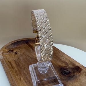May include: A beaded bracelet with a cream and gold color scheme. The bracelet is displayed on a clear acrylic stand, set on a wooden tray. The bracelet features a woven pattern with gold-colored metal clasps.
