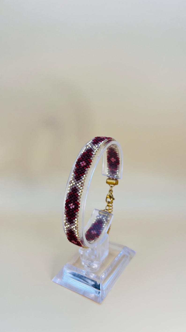 May include: A beaded bracelet with a geometric pattern in burgundy, white, and gold. The bracelet is displayed on a clear acrylic stand, showcasing its intricate design and craftsmanship. The clasp is gold-toned.