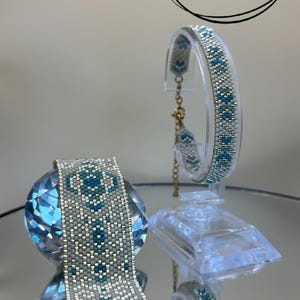 Puede incluir: Dos pulseras de cuentas con un patrón geométrico en verde azulado y plata. Una pulsera se muestra plana, la otra sobre un soporte transparente. Las pulseras están sobre una superficie reflectante. La marca "Miv Sieradenstudio" está en el fondo.