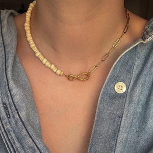 May include: A necklace featuring a combination of cream-colored beads and a gold-toned chain. The necklace has an infinity symbol pendant and is worn on a denim shirt.