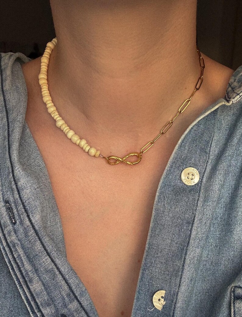 May include: A necklace featuring a combination of cream-colored beads and a gold-toned chain. The necklace has an infinity symbol pendant and is worn on a denim shirt.