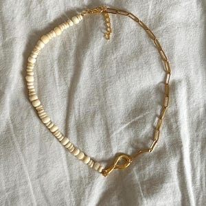 May include: A gold-toned necklace with a unique design. One half features small, off-white beads, while the other half is a gold chain. The necklace is connected by a gold clasp. The jewelry is displayed on a white surface.