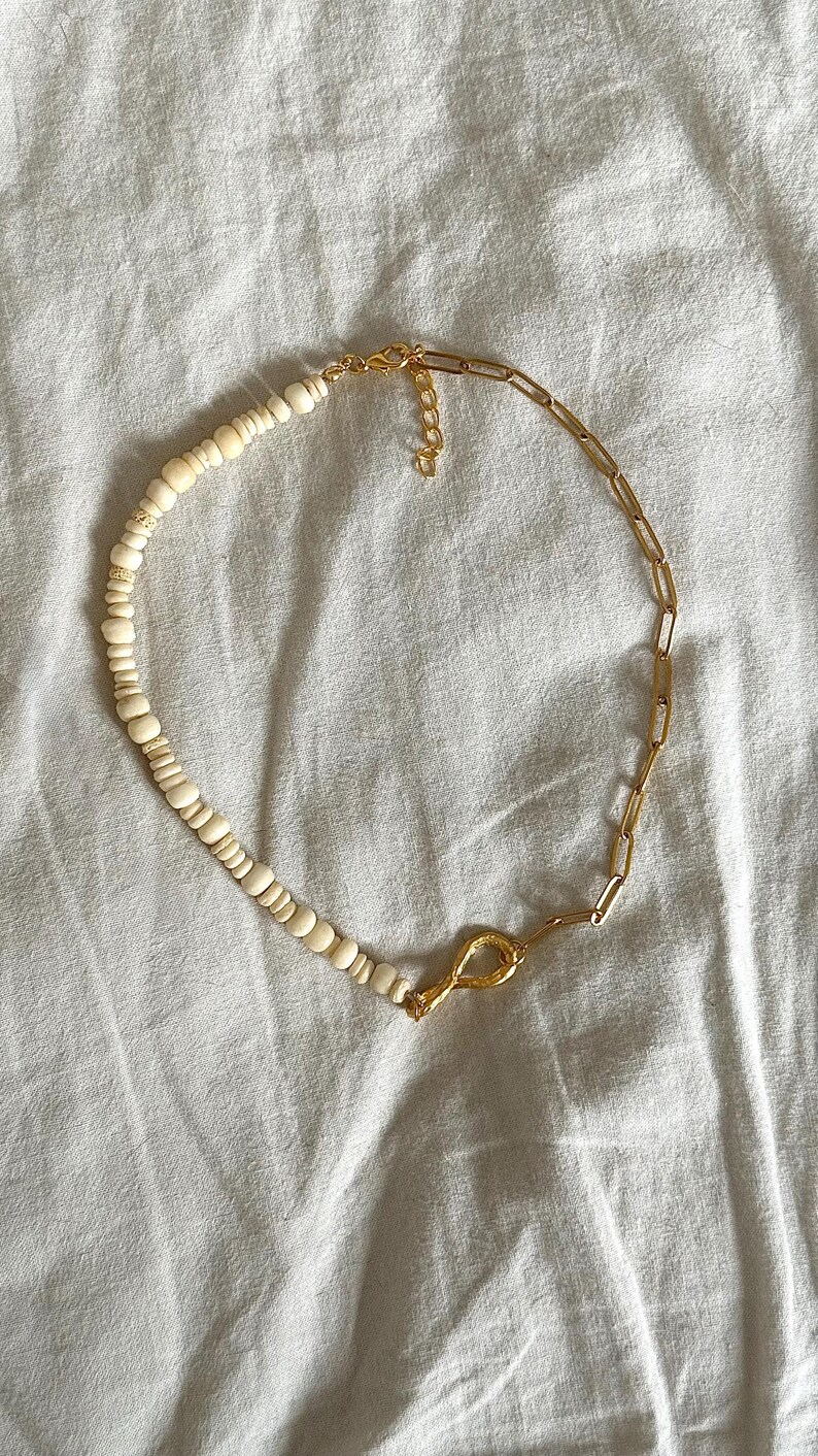 May include: A gold-toned necklace with a unique design. One half features small, off-white beads, while the other half is a gold chain. The necklace is connected by a gold clasp. The jewelry is displayed on a white surface.