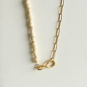 May include: A necklace featuring a combination of cream-colored, round beads and a gold-colored chain. The chain is connected to the beaded section with a gold-colored infinity symbol clasp. The necklace is displayed on a white surface.
