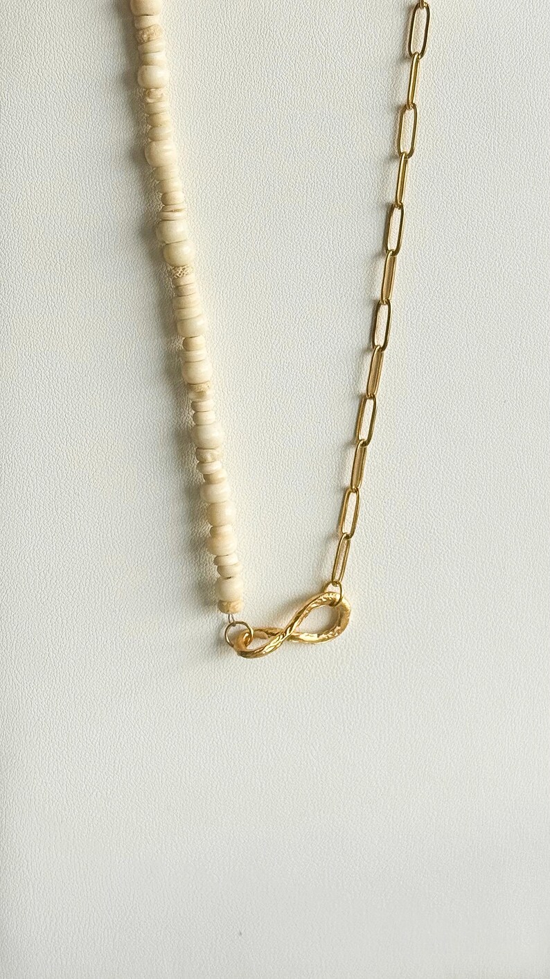May include: A necklace featuring a combination of cream-colored, round beads and a gold-colored chain. The chain is connected to the beaded section with a gold-colored infinity symbol clasp. The necklace is displayed on a white surface.