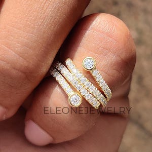 Può includere: Un anello dorato con un design a spirale, caratterizzato da più file di piccole pietre scintillanti. Due pietre più grandi, incastonate a castone, sono poste alle estremità della spirale. L'anello è sul dito di una persona, con il marchio "ELEONEJEWELRY" visibile.