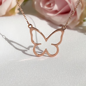 Op de afbeelding: Rosegouden vlinderhanger ketting. De delicate ketting heeft een vlindervormige hanger die aan een fijne ketting hangt. De achtergrond bevat roze rozen en witte bloemen, wat een zachte, romantische esthetiek creëert. De ketting wordt op een wit oppervlak weergegeven.