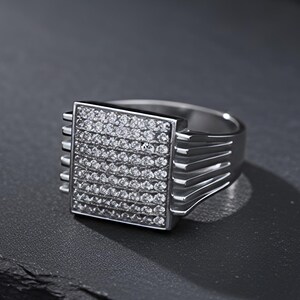 Può includere: Un anello d'argento con una faccia quadrata ricoperta di piccole pietre trasparenti. L'anello ha una fascia spessa e un design strutturato sui lati della faccia quadrata. Presentato su una superficie scura e strutturata.
