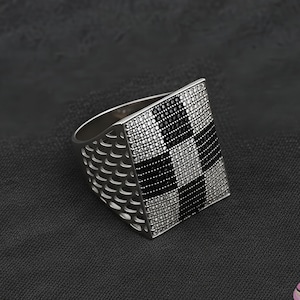 May include: A silver ring with a large, square face. The face features a checkerboard pattern of black and clear stones. The band has a textured design. The ring is set against a dark background.