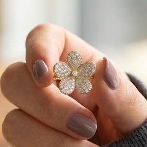 May include: A gold ring featuring a five-petaled flower design, with each petal densely encrusted with small, clear, sparkling stones. The center of the flower is marked by a single, slightly larger, clear stone, creating a bright, reflective appearance.