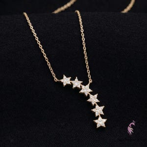 May include: A gold-colored necklace with a chain and six star-shaped pendants. The stars are encrusted with small, clear gemstones. The necklace is displayed against a black background.