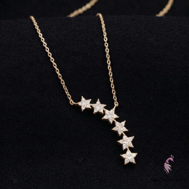 May include: A gold-colored necklace with a chain and six star-shaped pendants. The stars are encrusted with small, clear gemstones. The necklace is displayed against a black background.