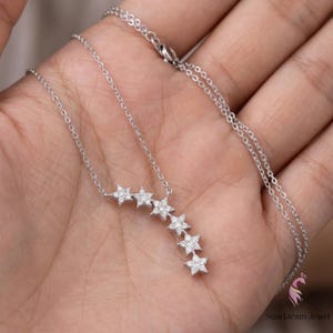 May include: A silver necklace featuring a series of star-shaped pendants, each encrusted with small, sparkling stones. The necklace is displayed against a neutral background, highlighting the delicate design and craftsmanship of the jewelry.