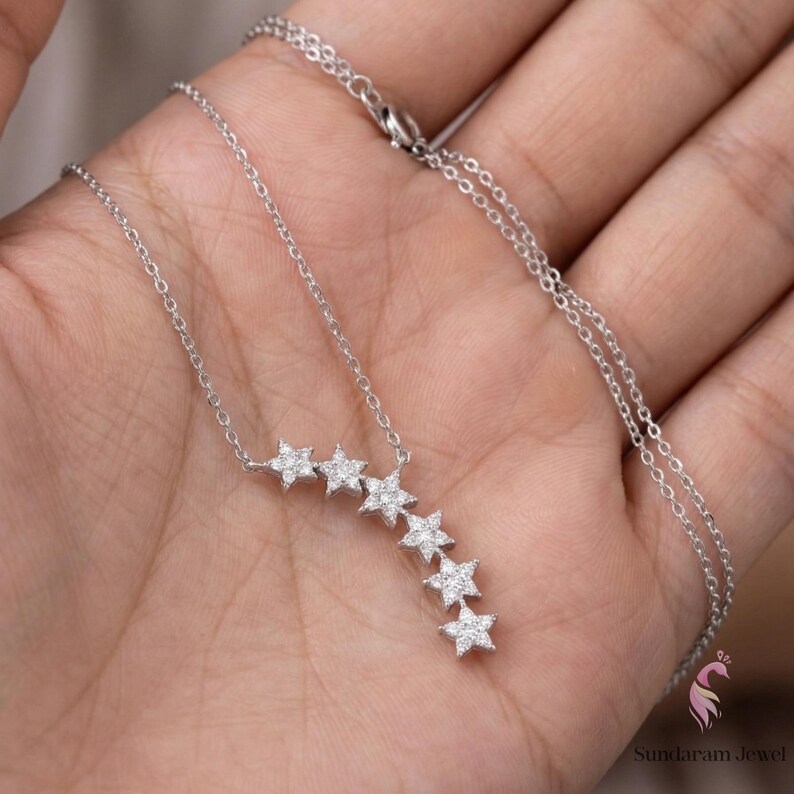 May include: A silver necklace featuring a series of star-shaped pendants, each encrusted with small, sparkling stones. The necklace is displayed against a neutral background, highlighting the delicate design and craftsmanship of the jewelry.