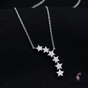 May include: A silver necklace with a series of star-shaped pendants. The stars are arranged in a cascading pattern, with the chain visible against a black background. The necklace is a delicate piece of jewelry.