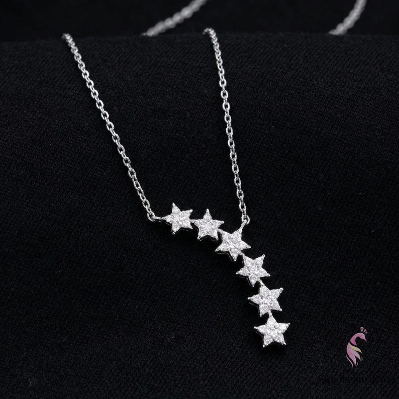 May include: A silver necklace with a series of star-shaped pendants. The stars are arranged in a cascading pattern, with the chain visible against a black background. The necklace is a delicate piece of jewelry.