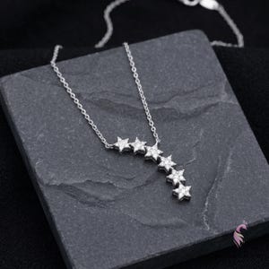 May include: A silver necklace featuring a cascade of six star-shaped pendants, each encrusted with small, sparkling gemstones. The necklace is displayed on a dark gray, textured surface, with a black background.