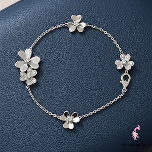May include: A silver bracelet with flower-shaped charms. Each charm has a central cluster of small, sparkling stones. The bracelet is connected by a delicate chain and has a clasp closure. The background is a solid, dark blue.