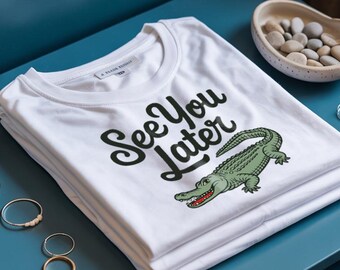 Puede incluir: Camiseta blanca con la frase "See You Later" y una ilustración de un cocodrilo. El cocodrilo es verde con la boca roja. La camiseta está doblada sobre una superficie azul.