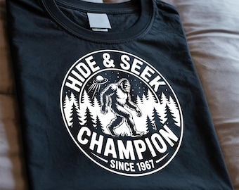 May include: Black t-shirt featuring a white circular graphic with the text "HIDE & SEEK CHAMPION SINCE 1967". The graphic includes a Bigfoot illustration, a UFO, and a forest silhouette.