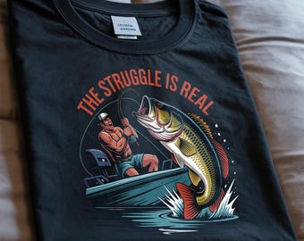 Puede incluir: Camiseta negra con un gráfico de un pescador en un barco luchando contra un pez grande. El texto "THE STRUGGLE IS REAL" está en naranja sobre la ilustración. La camiseta está hecha de un material suave.