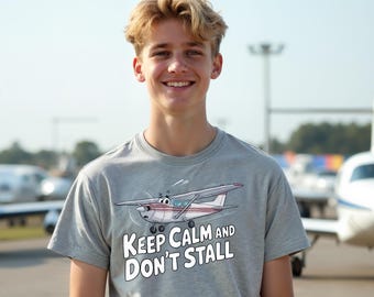 May include: A gray t-shirt featuring a cartoon airplane with the text "Keep Calm and Don't Stall." The shirt is worn by a person with blond hair, smiling in front of an airport background.