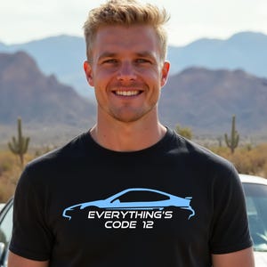 Könnte beinhalten: Schwarzes T-Shirt mit hellblauer Auto-Umrisszeichnung und dem Text "EVERYTHING'S CODE 12". Die Person lächelt vor einem Auto und einer Wüstenlandschaft.
