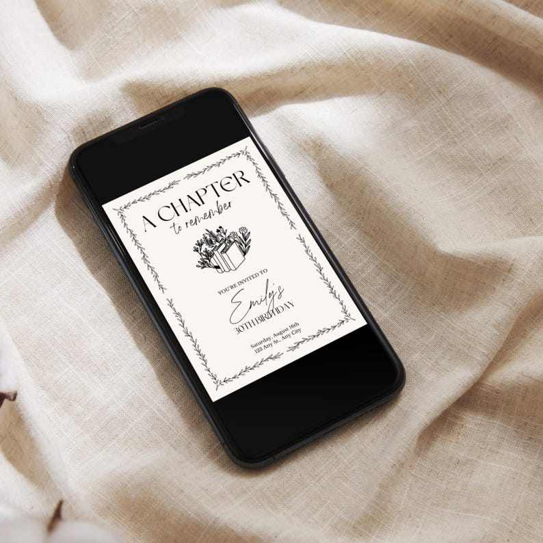 May include: A black smartphone displays a digital invitation with the text "A Chapter to Remember." The invitation features a floral border and the words "Emily's 30th Birthday." The phone rests on a beige fabric surface.