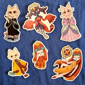 May include: A collection of six colorful stickers featuring cartoon characters with cat-like features. The characters are dressed in various outfits, including robes and dresses, and some are holding objects like a flute and a circular emblem. The stickers are on a blue background.