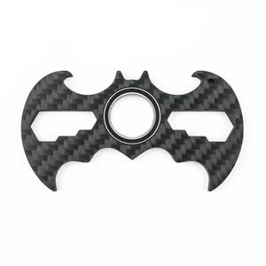 May include: A black carbon fiber multi-tool in the shape of a bat symbol. The tool has a central circular opening with a silver ring, and several hexagonal and curved cutouts. The surface has a textured, woven pattern.
