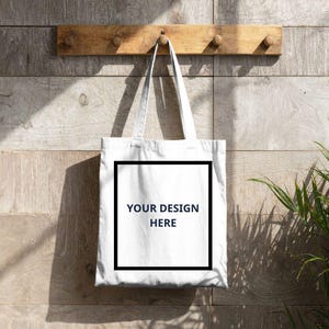 Puede incluir: Una bolsa de tela blanca con un borde cuadrado negro y el texto "YOUR DESIGN HERE" impreso en negro en el centro. La bolsa tiene dos asas largas para el hombro y cuelga de un gancho de madera en una pared de madera clara.