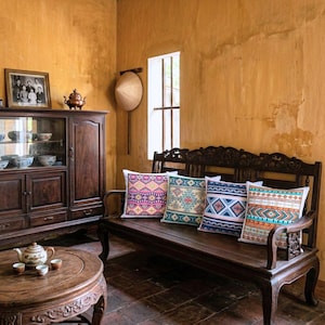 Puede incluir: Un banco de madera oscura con cuatro cojines decorativos con patrones geométricos coloridos. Los cojines están colocados en el banco frente a una ventana. La habitación tiene una estética vintage.