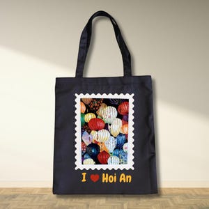 Puede incluir: Bolso tote negro con una colorida ilustración de faroles de Hoi An dentro de un marco de sello postal. El texto "I ❤️ Hoi An" está impreso en amarillo. El bolso tiene asas negras.