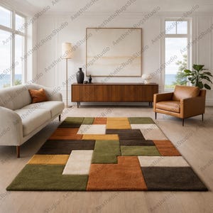 Pode incluir: Um tapete retangular com um padrão geométrico em tons de verde, castanho, laranja e creme. O tapete está colocado sobre um chão de madeira clara numa sala de estar. A sala também apresenta um sofá branco, uma poltrona de couro castanho e um móvel de madeira.