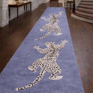 Op de afbeelding: Een lange, rechthoekige blauwe vloerkleed met twee luipaardontwerpen in zwart en wit. Het vloerkleed ligt in een gang met een donkere houten vloer en witte muren. De luipaardfiguren staan in een speelse pose.