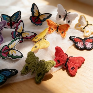 May include: A collection of embroidered butterfly patches in various colors, including blue, white, yellow, orange, red, and green. The patches are arranged on a white fabric, showcasing the detailed stitching and vibrant hues of each butterfly design.