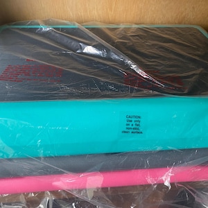 May include: A rectangular aerobic step platform featuring a teal base and a black textured top surface, encased in clear plastic packaging. A small white sticker on the side reads: CAUTION: Use only on a flat, non-skid, clean surface.