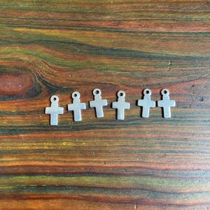 May include: Six small, silver-coloured cross charms with a loop at the top for stringing. The crosses are arranged in a row on a wooden surface with a warm, brown and green grain pattern.