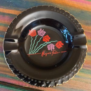 May include: Black ashtray with a gold rim and a floral design in red and purple. The text "Niagara Falls CANADA" is written in red cursive. The ashtray has three grooves for resting cigarettes.