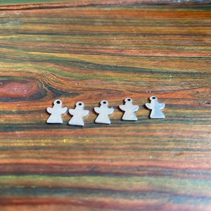 May include: Five small, silver-coloured angel charms with a hole at the top for stringing. The charms are arranged on a wooden surface with a warm, natural wood grain pattern. These charms are suitable for jewellery making or craft projects.