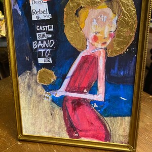 May include: Framed mixed media artwork featuring a figure in a red dress with a gold halo. The background is a mix of blue and gold. Text in the upper left corner reads "Stand Deep Rebel Wild CAST IN SUN BAND TO AIR."