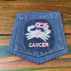 May include: A blue denim patch with a Cancer zodiac design. The patch features a white crab illustration with pink accents and the word "CANCER" in pink lettering. The patch has a frayed edge.