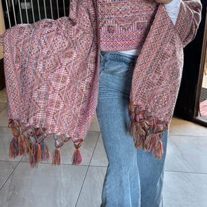 Peut inclure: Un châle rose tissé avec un motif géométrique et des pompons colorés. Le châle est drapé sur un haut blanc et un jean bleu. Les manches sont longues et évasées, et les pompons sont sur le bord inférieur.