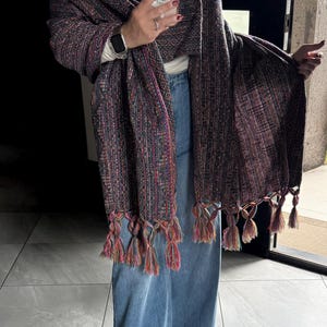 May include: A rectangular woven shawl featuring a dark, multi-colored textured pattern with vertical stripes of blue, red, and orange threads. The shawl is finished with long, knotted fringe tassels at the bottom edge, incorporating vibrant pink, orange, and blue yarn.