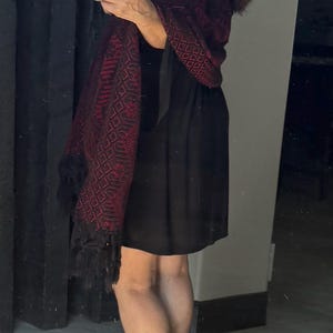May include: A black sleeveless dress paired with a dark red and black patterned shawl featuring geometric designs and black fringe trim. The ensemble is completed with black high-heeled pumps, creating a coordinated look.