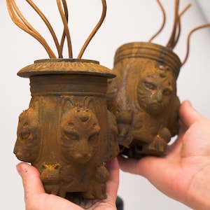 Peut inclure: Deux vases en métal rouillé avec des motifs de visages de chats. Chaque vase a un dessus évasé et un corps cylindrique orné de multiples visages de chats. Les vases ont une esthétique vintage et sont tenus dans les mains.